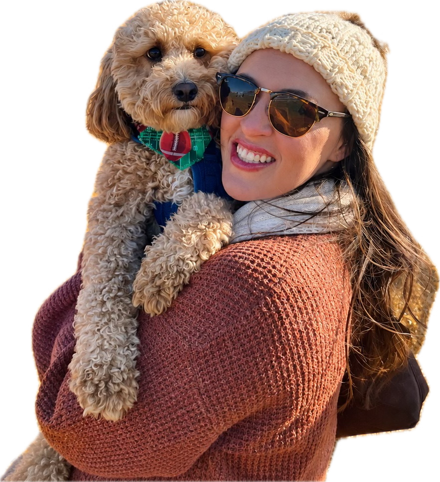 A smiling woman in sunglasses and a cream knit headband holding her curly golden doodle, who wears a green football-print bandana
